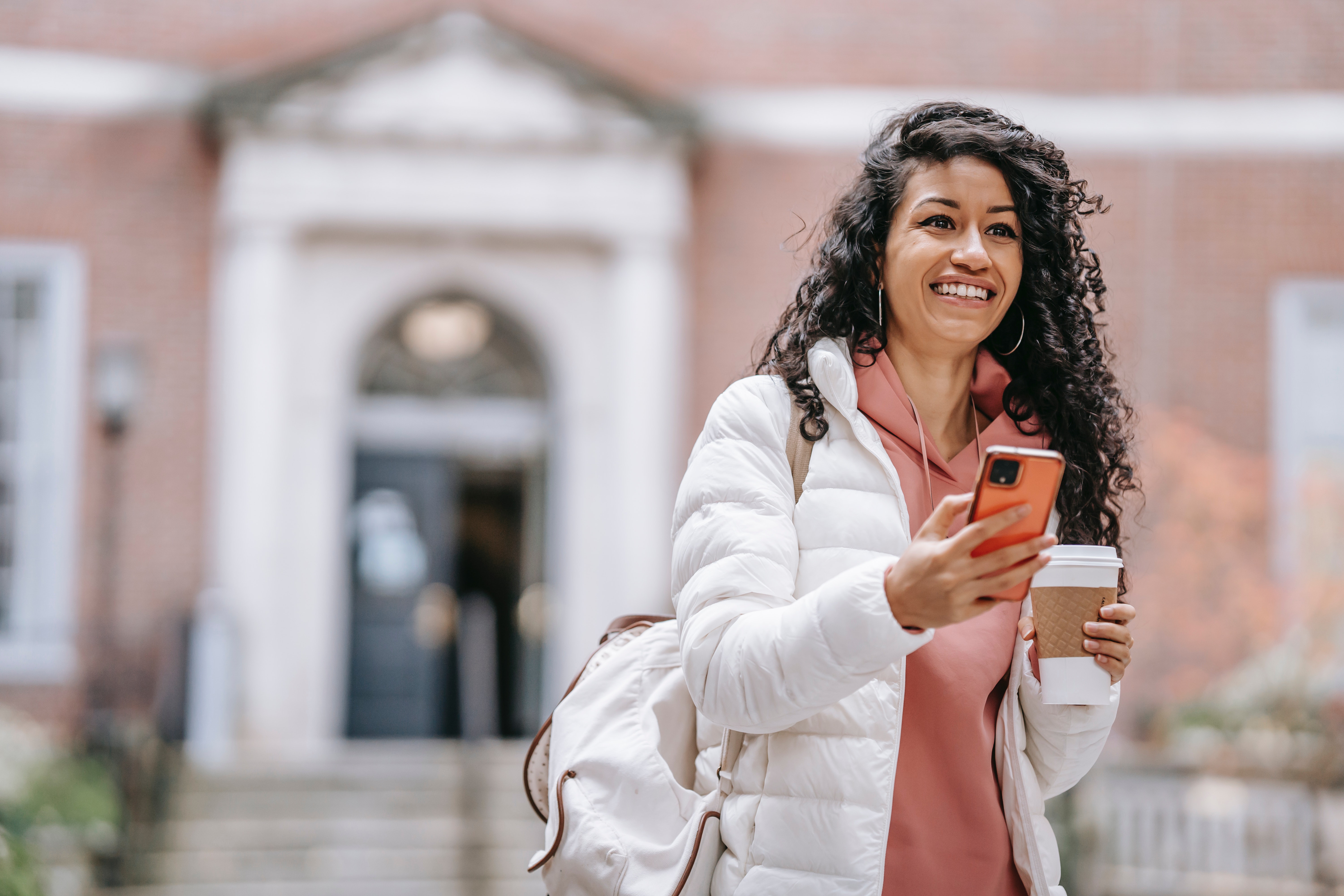 Smiling female college student holding a cup of coffee and a smartphone.